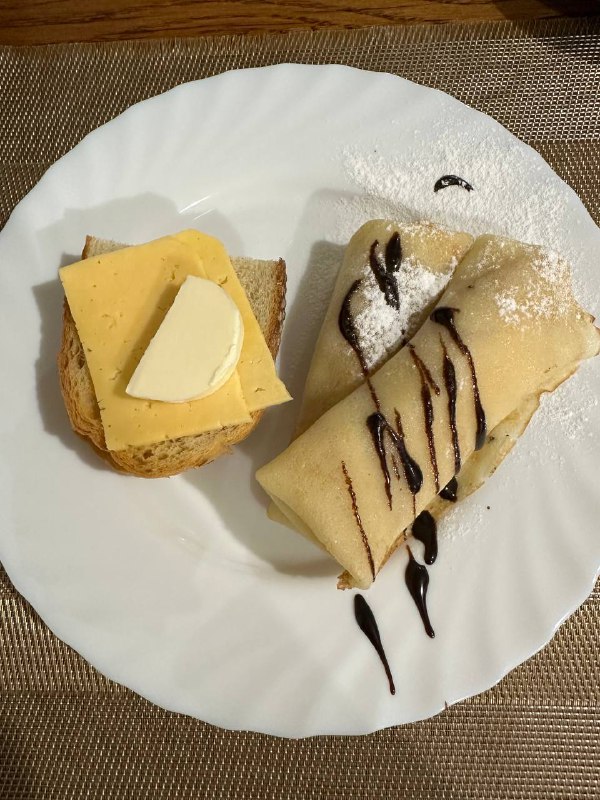 Slice Of Bread With Cheese And Butter, Alongside A Crepe Topped With Chocolate Sauce And Powdered Sugar