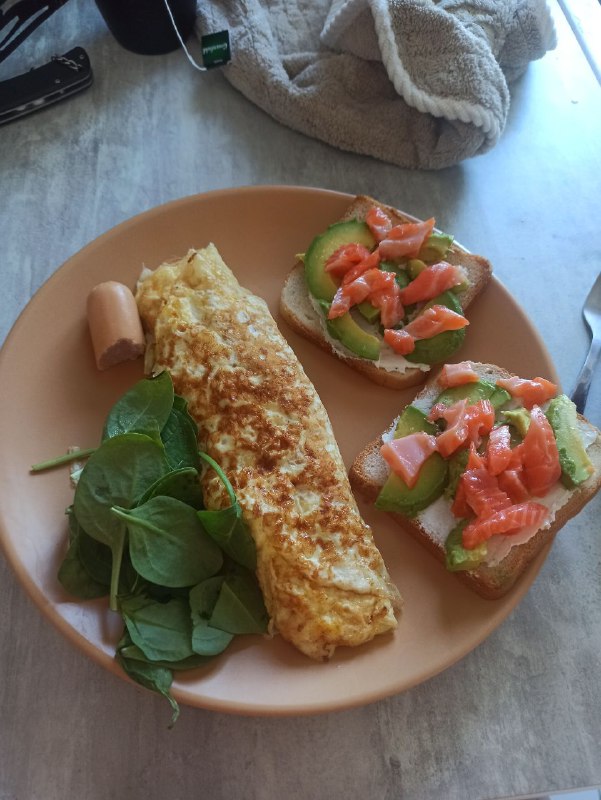 Combination Of Omelette, Smoked Salmon On Toast With Cream Cheese And Avocado, Spinach, And A Hot Dog Sausage